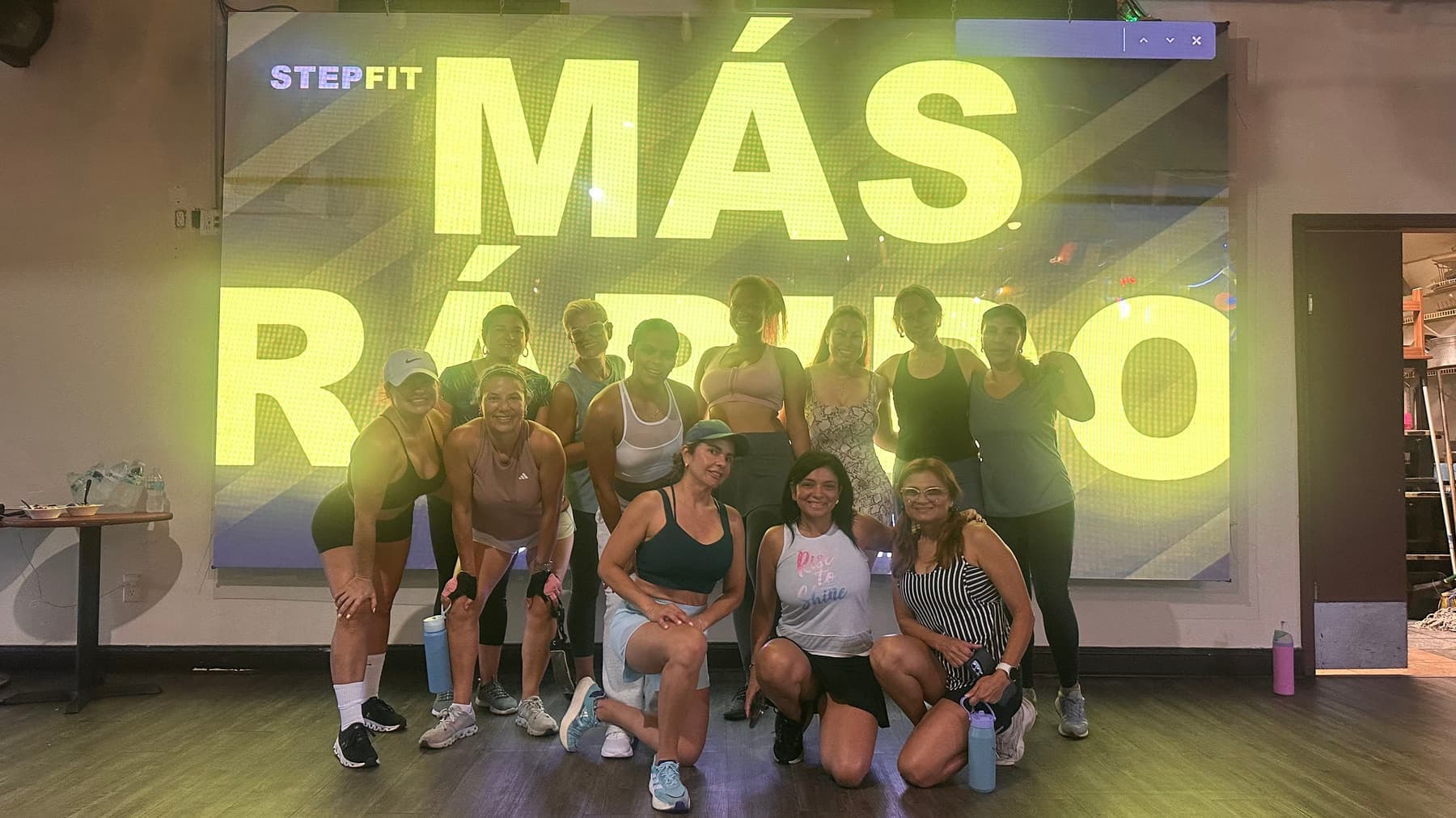 A group kneeling and standing in front of a wall-sized projection reading MÁS RÁPIDO, water bottles on the floor beside them.
