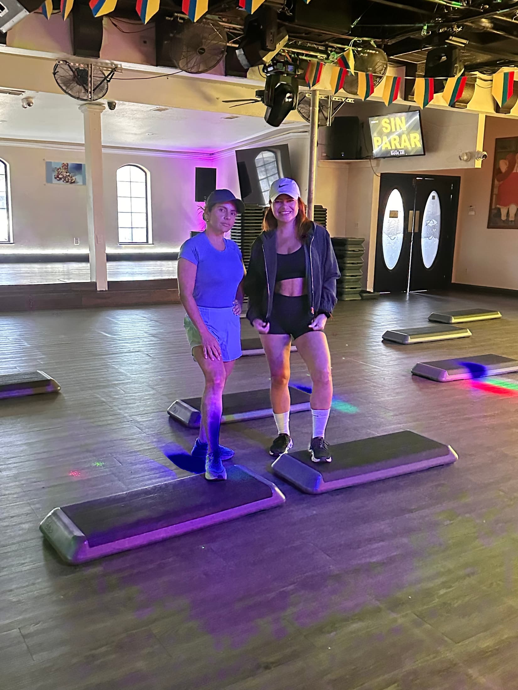Two people standing on step platforms in a bright hall before class, with SIN PARAR on a screen on the far wall.