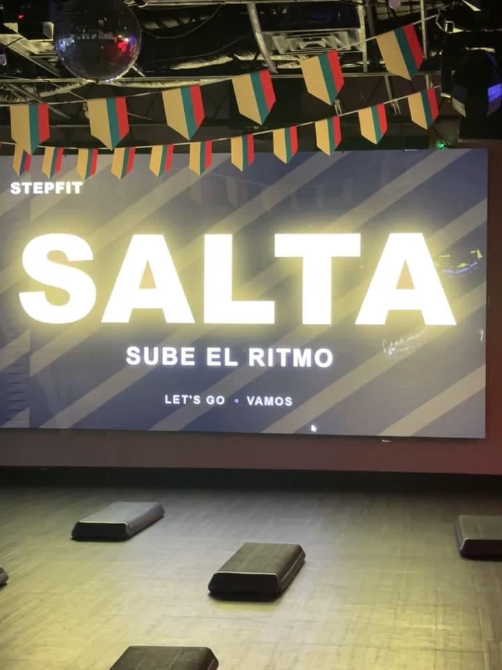 Step platforms spaced out across an empty wooden floor in front of a wall-sized projection reading SALTA, SUBE EL RITMO, with paper bunting and a mirror ball overhead.