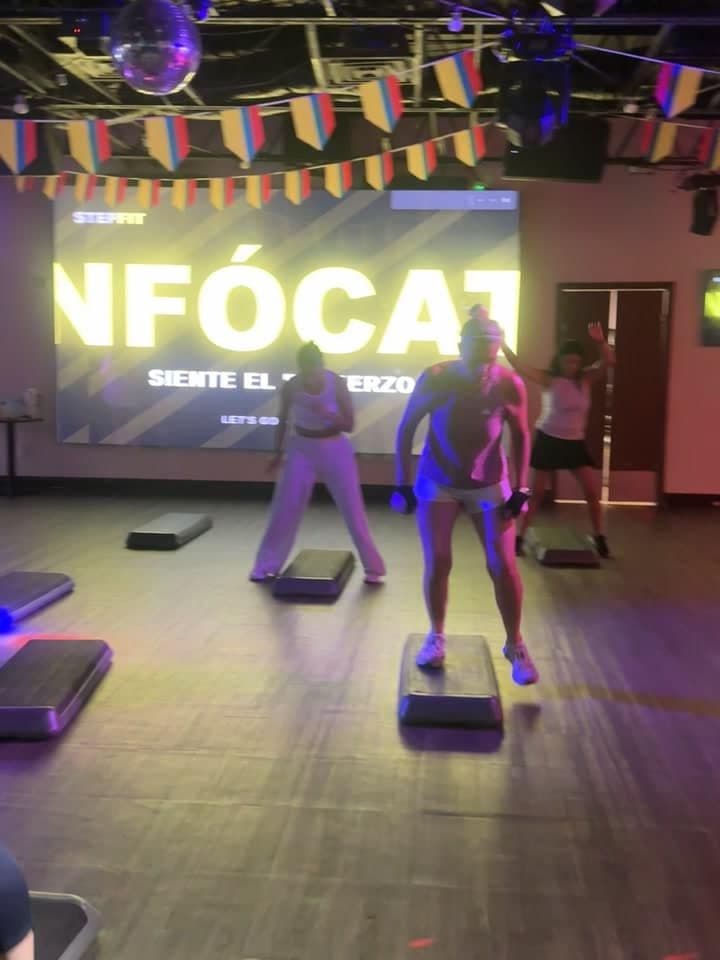 Three people stepping up onto platforms in a room lit magenta and yellow, the word ENFÓCATE projected on the screen behind them.