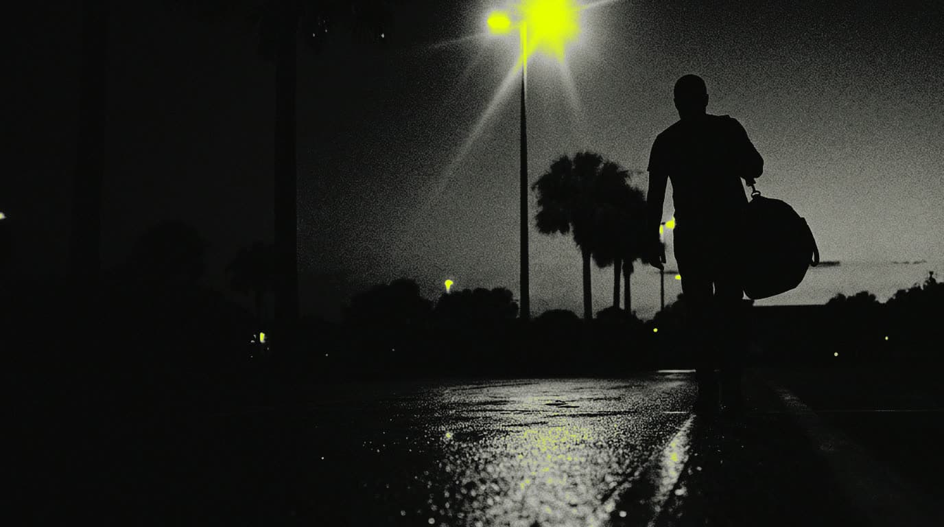 A single figure in silhouette walking away across an empty parking lot at dusk, a bag over one shoulder, palms and one lot lamp above