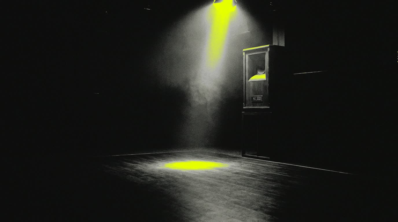 A tall speaker cabinet standing at the edge of an empty wooden dance floor, lit by one narrow beam from the rig overhead